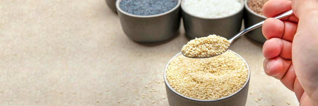 White sesame seeds, sesame seeds in a black saucer on a background of various seeds. Male hand sprinkles sesame seeds with a spoon.