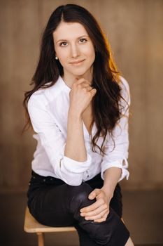 Portrait of a happy young brunette woman in a white shirt sitting on a chair.