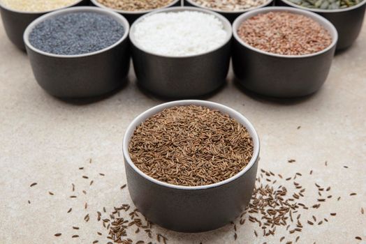 Caraway seeds. Cup with caraway seeds on a background of various seeds in black containers on a marble table top.