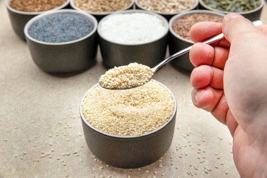 White sesame seeds, sesame seeds in a black saucer on a background of various seeds. Male hand sprinkles sesame seeds with a spoon.