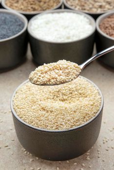 White sesame seeds, sesame seeds in a black saucer on a background of various seeds. Male hand sprinkles sesame seeds with a spoon.