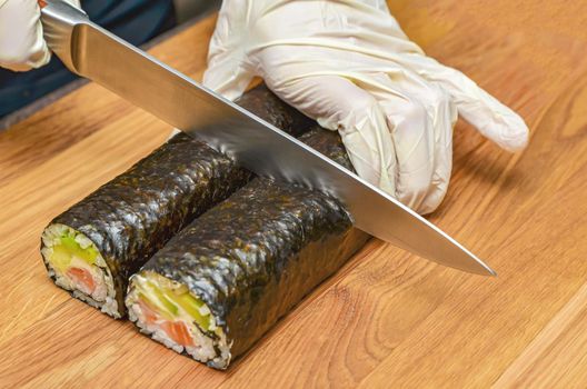 The process of making Japanese sushi. Knife in hand cuts a roll close-up on a wooden board. Female hands in rubber gloves