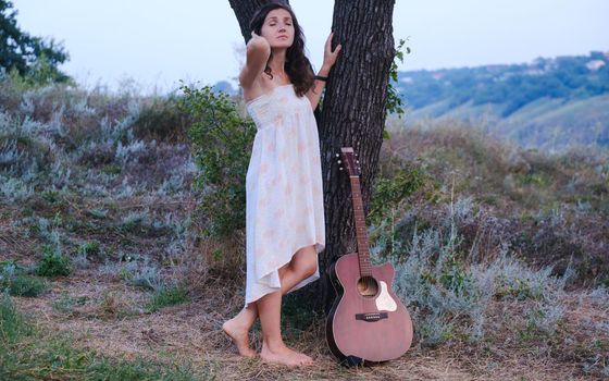 Beautiful romantic girl with dark hair and white dress with guitar outdoor. Photo of sensual woman. Art work. Music background.