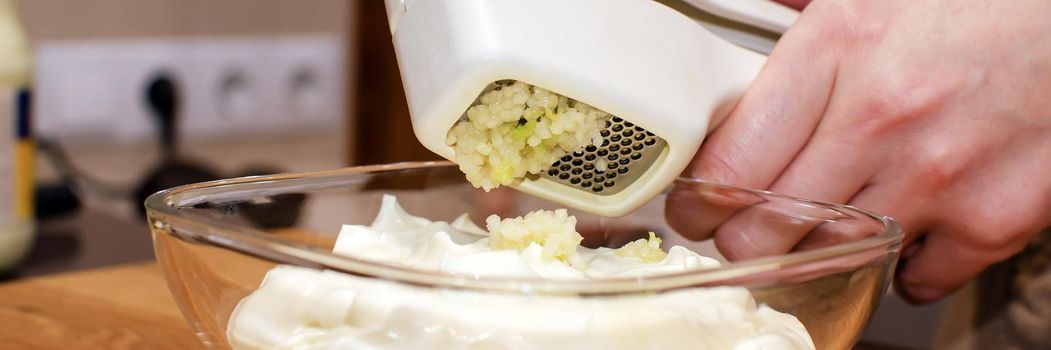 Garlic press made of stainless steel and white plastic. Making garlic sauce in a glass saucer.