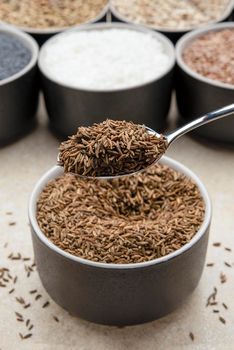 Caraway seeds. Spoon with caraway seeds in hand against a background of various seeds in black containers on a marble table top.