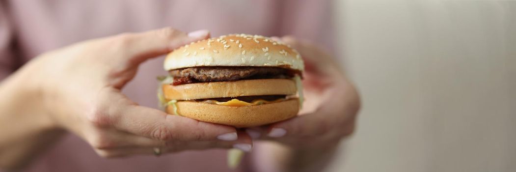 Close-up of female holding tasty burger, soft bun with meat cheese sauce and vegetables. Fast food, unhealthy, takeaway, order delivery, hungry concept. Blurred background