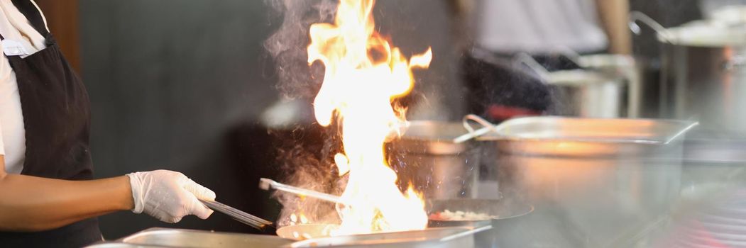 Close-up of work of cook in kitchen, preparing meal at open fire grill, fire in chefs pan, fresh cooked dish. Cooking, street food, takeaway, improvisation, show concept. Blurred background