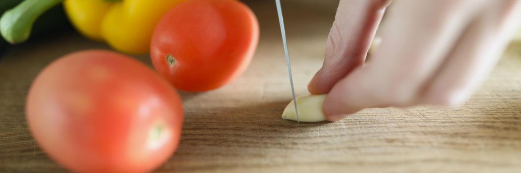 Close-up of person cutting garlic with sharp knife, fresh vegetables on wooden cutting board. Ripe tomatoes, yellow pepper for salad. Cooking, chef, food, kitchen concept