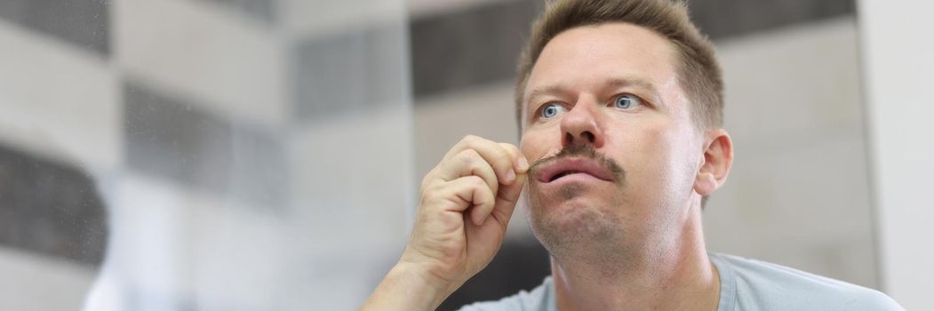 Portrait of man pull his mustache with hand, fake mustache, no beard. Reflection in mirror, start day with beauty procedure, take care of appearance. Bathroom, face care, get ready concept