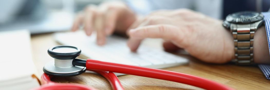 Close-up of medical equipment red stethoscope on workplace. Doctor typing on keyboard, medical building for health checkup. Medicine, clinic, appointment, healthcare concept