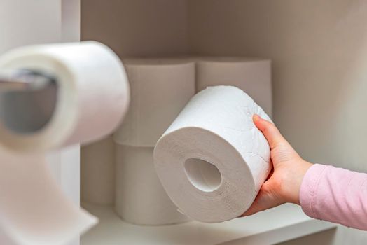 Roll of white toilet paper in hand. The child's hand takes a roll of toilet paper from the cabinet. Lots of toilet paper on background.