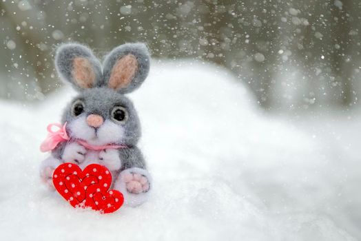 Happy Valentine's Day greeting card or banner. Plush rabbit holding a red heart symbol of love. Holidays Happy Valentine's Day. Snowfall on the background of the forest.