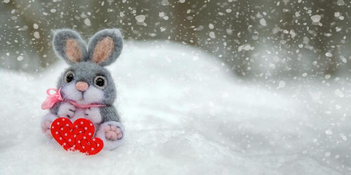 Happy Valentine's Day greeting card or banner. Plush rabbit holding a red heart ka symbol of love. Holidays Happy Valentine's Day. Snowfall on the background of the forest.