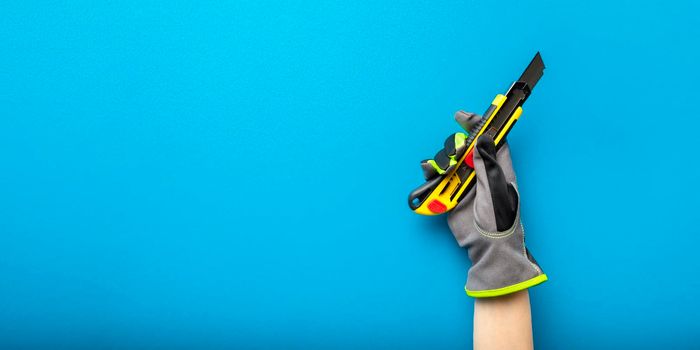 Construction knife. Stationery knife in hand with a glove on a blue background. Construction and renovation theme banner. Place for text or design.