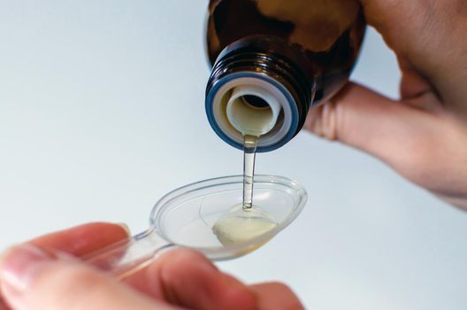 Close-up of female hands holding a bottle of expectorant cough syrup and pouring it with a measuring spoon. Health care and treatment concept. Blurred, white background.