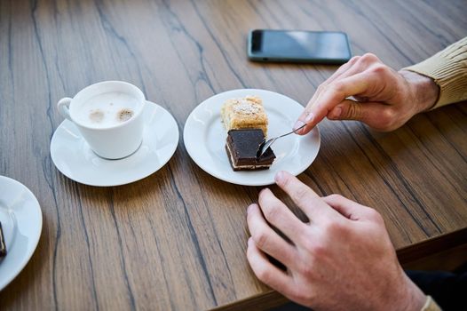 Cropped view of the hands of a man holding tea spoon and eating delicious baked chocolate cake, sitting at wooden table during coffee break. Smartphone with empty screen for copy ad space on table