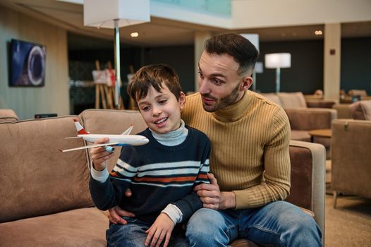 Handsome young loving father playing toy plane with his adorable son while relaxing in VIP lounge at international airport departure terminal and waiting to board flight during family travel