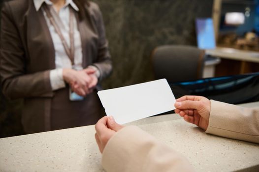 Focus on a white blank ticket with copy space for advertising text in the hands of unrecognizable woman standing by check-in counter in the VIP lounge of the international airport departure terminal