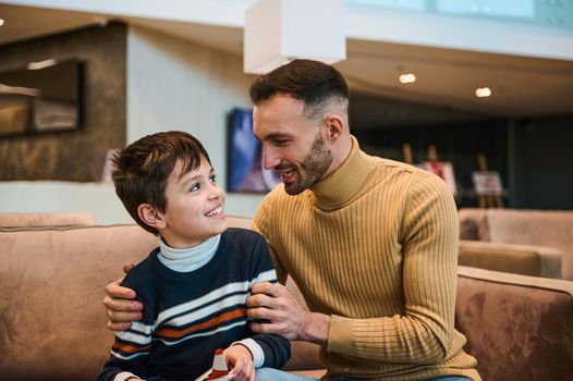 Handsome young dad communicating with his adorable son, sitting together on a comfortable couch in the VIP lounge of the airport departure terminal, waiting to board the flight. Family travel