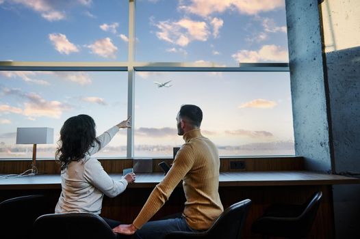 Rear view of a young married couple of passengers admiring a plane taking off, sitting at the panoramic windows in the departure terminal of an international airport, waiting to board the flight