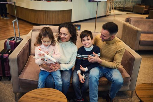 Overhead view of loving mom and dad tenderly hug their son and daughter playing with toy airplanes as they wait to board their flight in the VIP lounge of the international airport departure terminal.