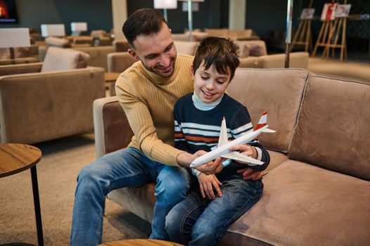 Handsome young Middle-Eastern man, loving father playing toys plane with his adorable son while resting in the VIP lounge in the international airport departure terminal during family travel
