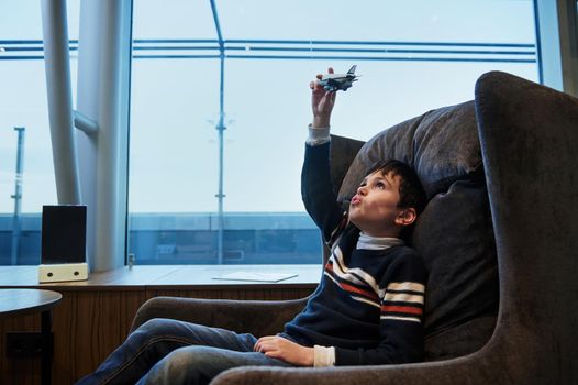 Adorable European teenage boy plays with toy airplane sitting by panoramic windows overlooking the runway at sunset while waiting to board flight at international airport departure terminal