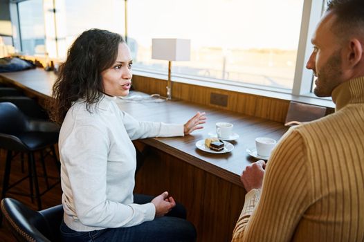 Focus on a Middle Eastern woman talking to her partner while having a delicious meal in the airport departures terminal dining room, seated by panoramic windows overlooking the runway.