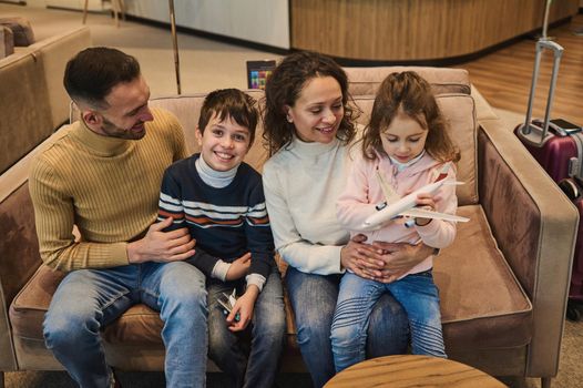 Happy multi-ethnic young loving mom and dad spending time together playing toy planes with their kids, son and daughter at the airport while waiting for a flight. Family Air travel concept