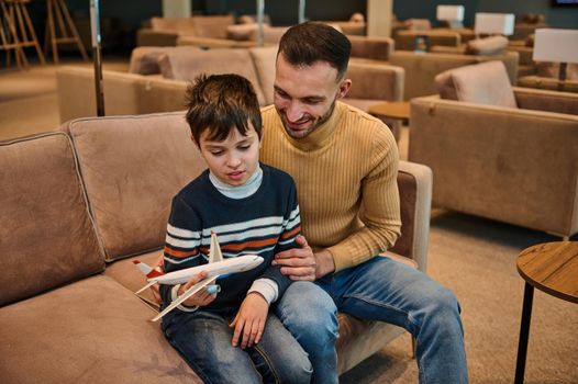 Handsome young Middle-Eastern man playing toys plane with his younger brother while resting in the VIP lounge in the international airport departure terminal during family travel