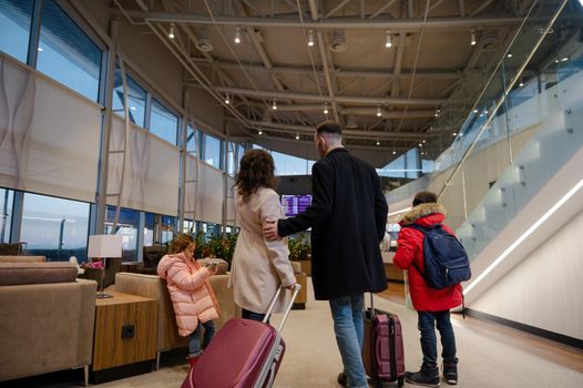 Rear view of traveling family in warm clothes with suitcases checking flight information on board with timetable, standing in the international airport departures terminal, waiting to board flight