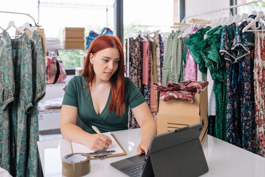 young entrepreneur, looking at her tablet and writing down the new orders for her online clothing shop. woman owner of a small online business. work and business concept.natural light from window, background with clothes racks and clothes.