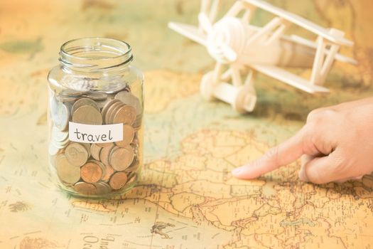 Hand pointing attractions in a map on a table with a glass jar with coins and travel inscriptions and a wooden toy plane nearby.