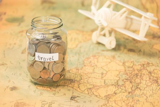 Glass jar with coins and travel inscription on world map with wooden toy plane on table. Travel concept and saving money.