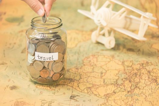 Hand putting coins in a clear jar on the table to save for travel. Glass jar with coins and an inscription travel on the world map with wooden toy plane on the table.