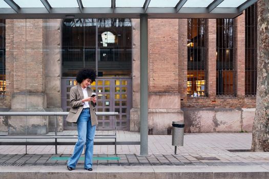 young latin woman waiting at the bus stop checking her mobile phone, concept of urban and modern lifestyle