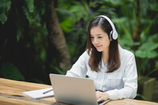 happy young asian woman smile using laptop and wearing white headphone while sitting at cafe. Young asian woman sitting in a coffee shop and video call conference on laptop.