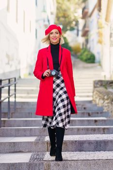 Full length of adult female with blond hair, in stylish outfit and beret smiling and looking at camera while walking on stairs on narrow street and photographing during holidays