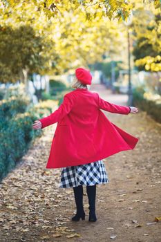 Full body back view of unrecognizable female in red outfit spinning on path with fallen leaves in park on autumn day