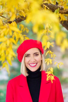 Confident excited young stylish woman with blond hair and makeup in stylish red coat and beret, smiling and looking at camera while standing near autumn tree with bright yellow leaves
