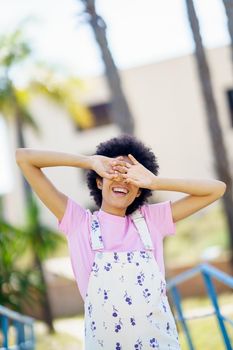 Content female in casual overall covering eyes with hands while standing on street with building against blurred background in city