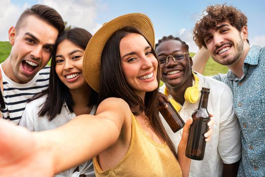 Happy multiracial group of friends take selfie while enjoying some beer outdoors in nature. Looking at camera. Friendship and technology concept.