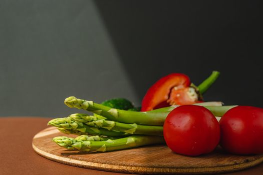 Vegetables lie on a wooden board: tomatoes, asparagus, cucumbers, red bell peppers. brown, dark gray background. place for text