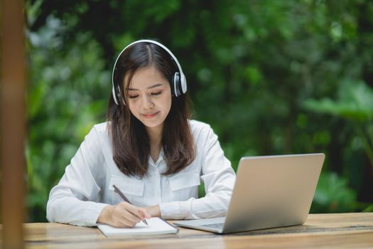 happy young asian woman smile using laptop, wearing white headphone and writing notebook. Young asian woman sitting in a coffee shop and video call conference on laptop.