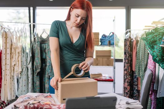 young business woman, closing a cardboard box with adhesive tape, order for customer of her online clothing shop. Young woman preparing a package for a client in the office. work and business concept. background with clothes racks and clothes.