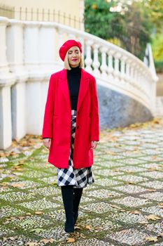 Full body of attractive female in red coat with beret standing on street with closed eyes near white fence in city