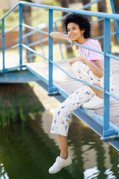Full body side view of cheerful African American female in casual outfit sitting on footbridge over lake while enjoying summer day in park