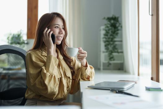 Corporate Communication. Asian Businesswoman Talking On Mobile Phone Working On Laptop In Modern Office