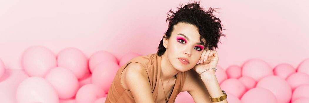 Stylish brunette with pink make-up on the background of many airy pink pastel balloons. Festive portrait in the studio. Web banner.