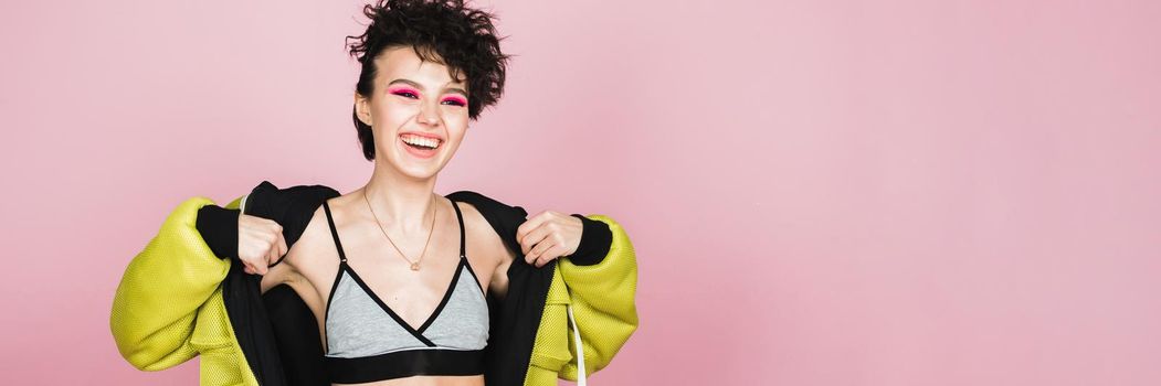 Beautiful teenager girl sincerely smiles and laughs in sports stylish clothes in the studio on a pink background. Web banner.